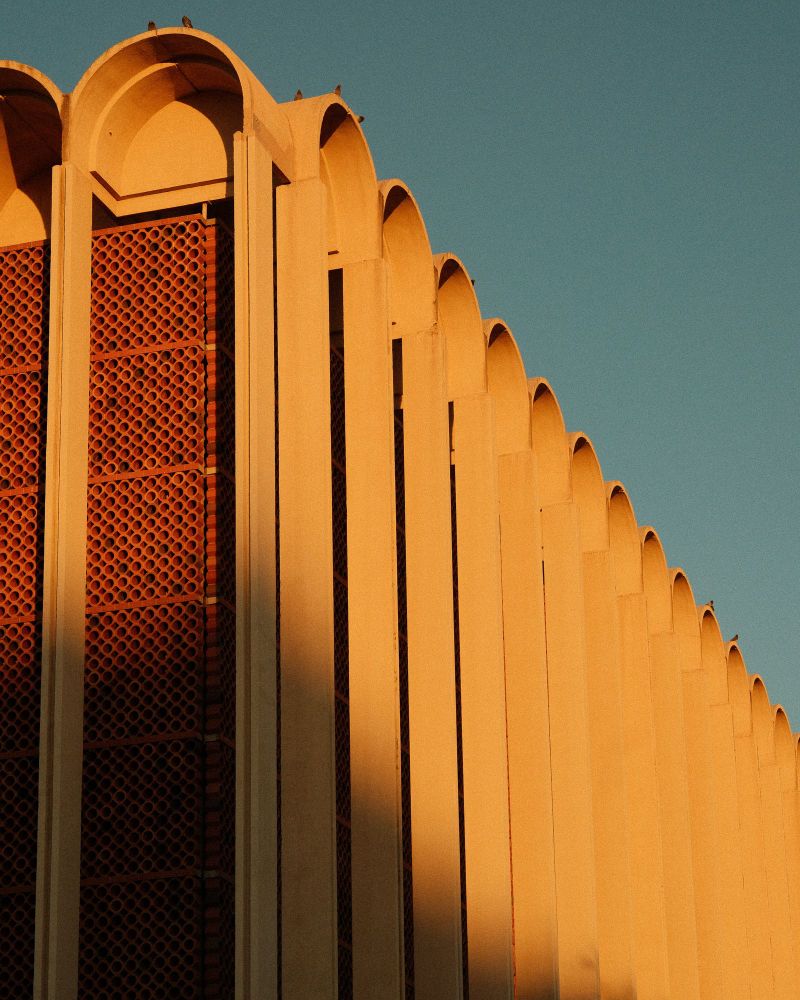 A corner view of a building that contains repeating arches as if a book laying with is spin into the air. The building is a warm white color from the sunset light hitting it. 