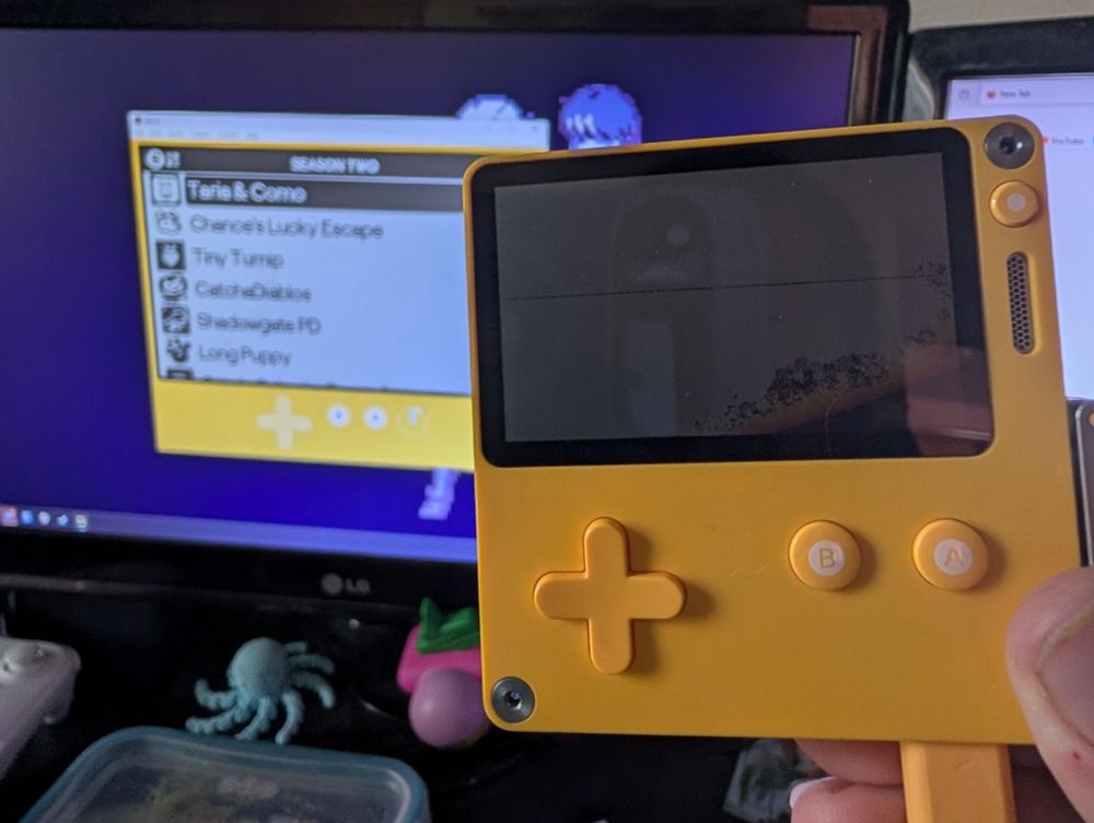 A Playdate console with a broken screen plugged into a computer where the Mirror app for the device allows it to be seen and heard on the computer instead.