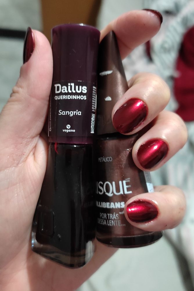 Foto de uma mão segurando dois esmaltes: "sangria" da marca Dailus (um vermelho transparente) e "por trás dessa lente" da Risqué (um marrom metalizado)
Nas unhas está a esmaltação que combina ambos os esmaltes, deixando o metalizado num tom vermelho escuro.