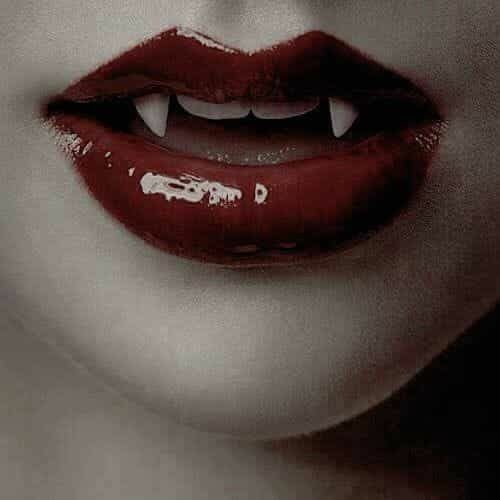 Close up of a mouth with vampire fangs peeking out. They’re wearing a dark red lipstick, glossed up, with a sharp ass Cupid’s bow (I love it)