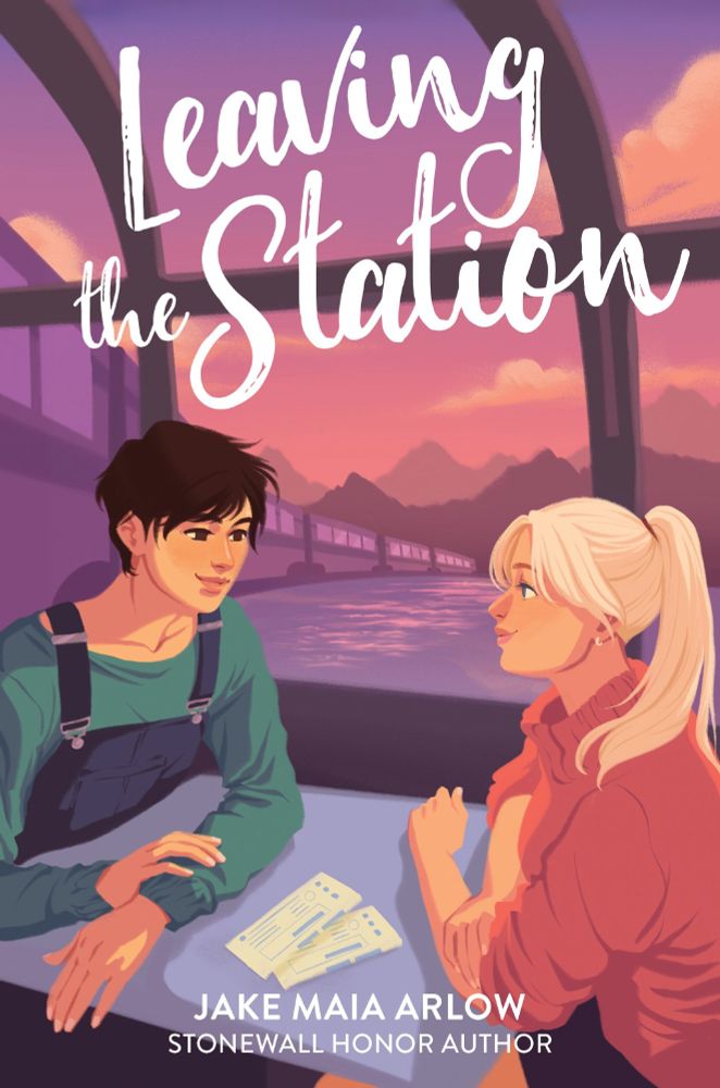Leaving the Station by Jake Maia Arlow (Stonewall Honor author)

Description: a young nonbinary person with short dark hair wearing a green longsleeve and dark blue overalls is seated across from a young blonde woman with a ponytail wearing a coral pink sweater. they are looking at each other smiling. they're both seated at a table together in the observation car of a train - out the window you can see the rest of the train cars trailing behind them, the mountains in the distance, and the pink-purple sunrise