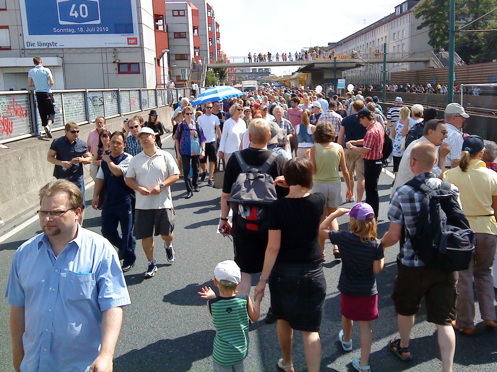 eine große menschenmenge spaziert dicht gedrängt über die abgesperrte autobahn 40. die stimmung wirkt fröhlich und entspannt. viele teilnehmende tragen sommerkleidung, einige kinder laufen an der hand ihrer eltern. links ist eine betonmauer mit graffiti, darüber ein großes blaues schild mit der aufschrift „40 – sonntag, 18. juli 2010 – die längste …“. rechts sind straßenbahngleise und gebäude zu sehen. im hintergrund überspannt eine fußgängerbrücke die szene. das bild zeigt einen abschnitt des still-leben ruhrschnellweg auf der a40.