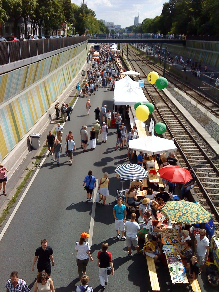 viele menschen spazieren gemütlich über die abgesperrte autobahn 40, die zwischen zwei bunten mauern verläuft. auf der rechten seite verläuft ein gleis, auf der straße stehen sitzbänke und sonnenschirme, an denen luftballons befestigt sind. über der szene spannt sich eine brücke, auf der weitere menschen stehen. es ist sonnig und wirkt wie ein sommerliches straßenfest. das foto wurde am 2010-07-18 aufgenommen, im rahmen des still-leben ruhrschnellweg auf der a40.