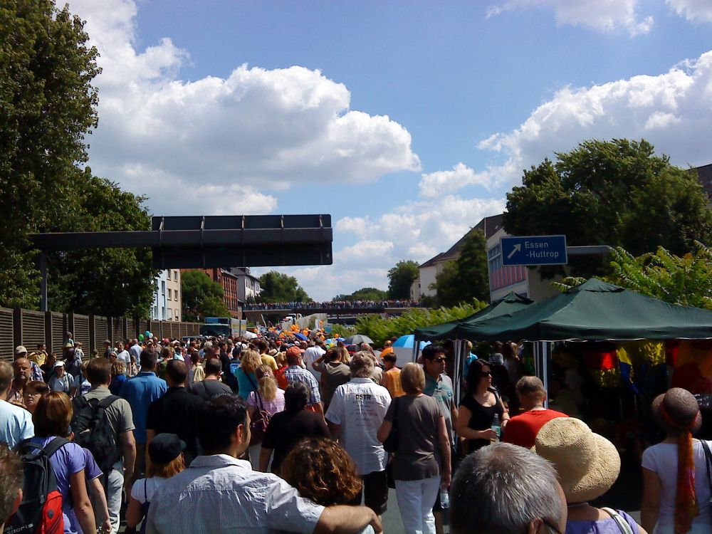 eine große menschenmenge spaziert dicht gedrängt über die abgesperrte autobahn 40. die stimmung wirkt fröhlich und entspannt. rechts im bild hängt ein schild mit der aufschrift 'essen huttrop'.
im hintergrund überquert eine brücke, auf der viele menschen stehen. das bild zeigt einen abschnitt des still-leben ruhrschnellweg auf der a40.