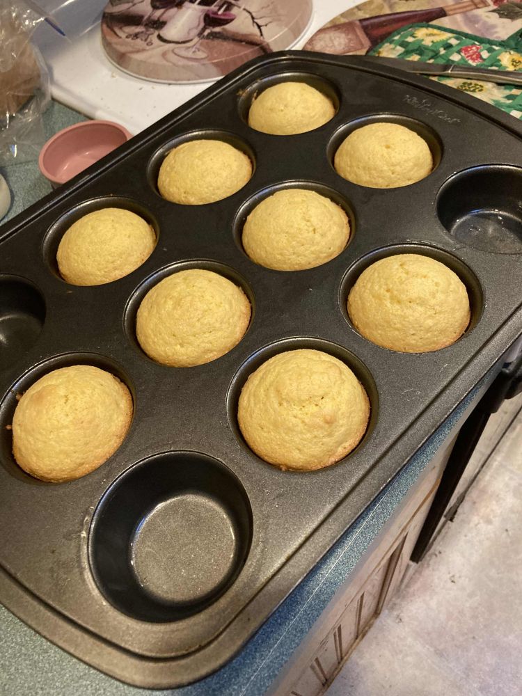 Cornbread muffins rest in a dark grey muffin tin. The muffins are a light golden brown and have only risen just above the lip of each muffin cup. The tray is stretched between a pale green countertop and the top of a white oven, and the shot is angled to show some of the off-white floor at the bottom right corner. On the oven, there are metal burner covers with scenes of painted still-lifes with wine. A green pot holder decorated with yellow flowers sits next to the muffin tray with a butter knife on top. In the top right corner, there is a loaf of bread wrapped in plastic and a pink measuring cup on the counter.