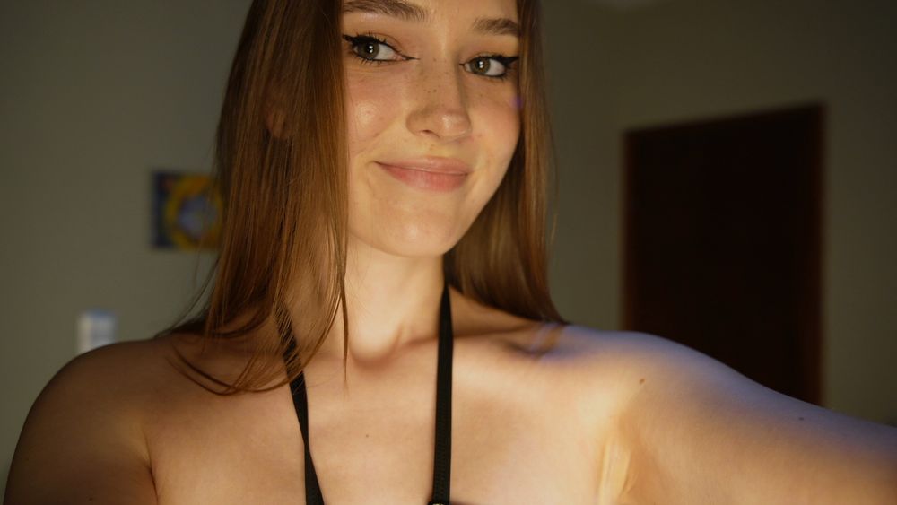 A young woman with long light brown hair is smiling softly at the camera. She is wearing a black halter top, her skin is softly illuminated by warm light. Her makeup is natural, with eyeliner accentuating her eyes. She looks confident and gentle.