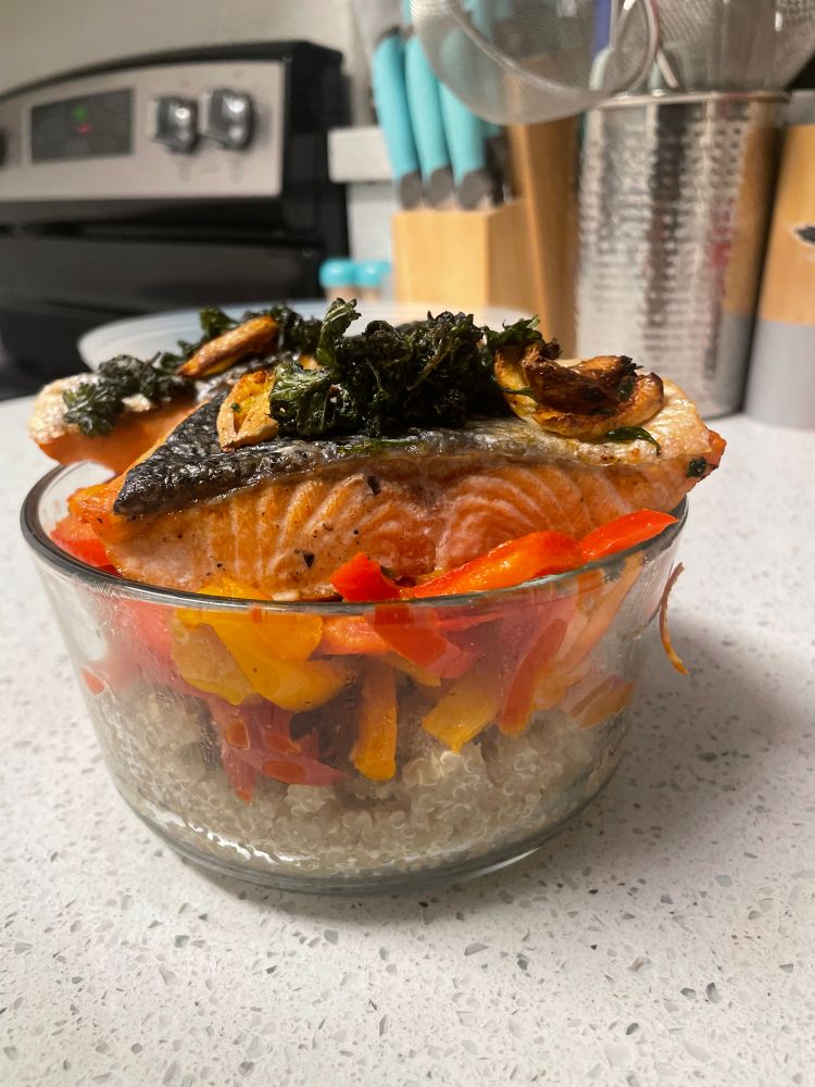 a picture of a bowl with quinoa, red and orange bell peppers and 2 pieces of salmon on top with crushed garlic and parsley (side profile)