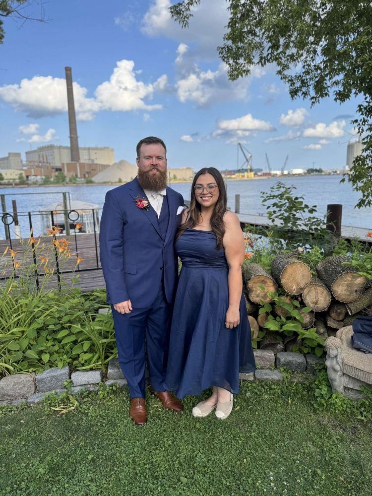 A man in a navy blue suit smiles next to a woman in a navy dress. They’re posing in front of greenery with a dock and lake behind them. 