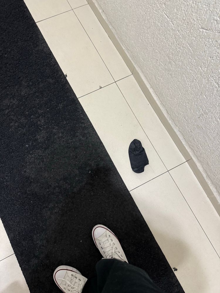 foto de um piso de cerâmica creme com carpete preto por cima. é possível ver os pés de alguém usando tênis branco e calça preta. no chão, está uma meia preta tipo soquete com sinais de sujeira. 