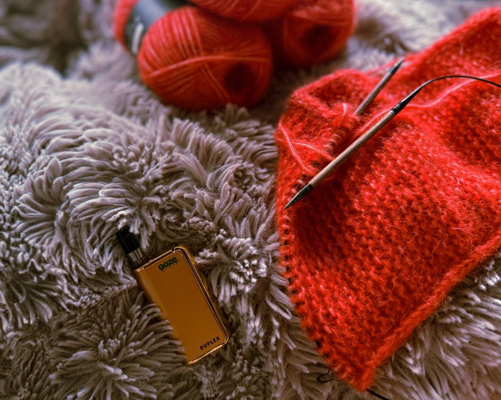gold vape sitting on fuzzy blanket next to red scarf in progress of being knit 