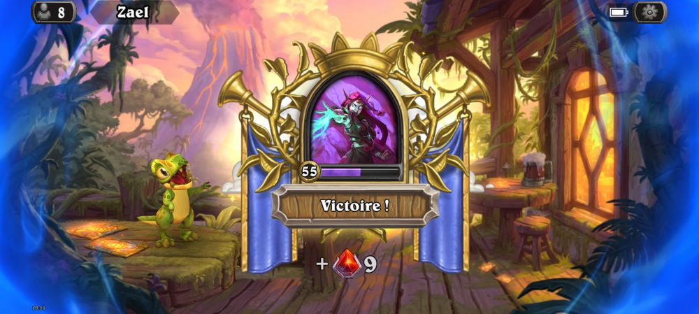 Écran de victoire Hearthstone contre 'Zael'. Un cadre doré orné entoure le portrait de l'adversaire vaincu. Le texte 'Victoire !' est affiché en français. En bas, une récompense de '+9' avec un symbole de gemme rouge est visible. Le familier Roi Krush apparaît à gauche dans un décor tropical avec de la végétation luxuriante typique de la jungle, un volcan en arrière plan et une hutte avec une chope de bière sur le côté droit.
