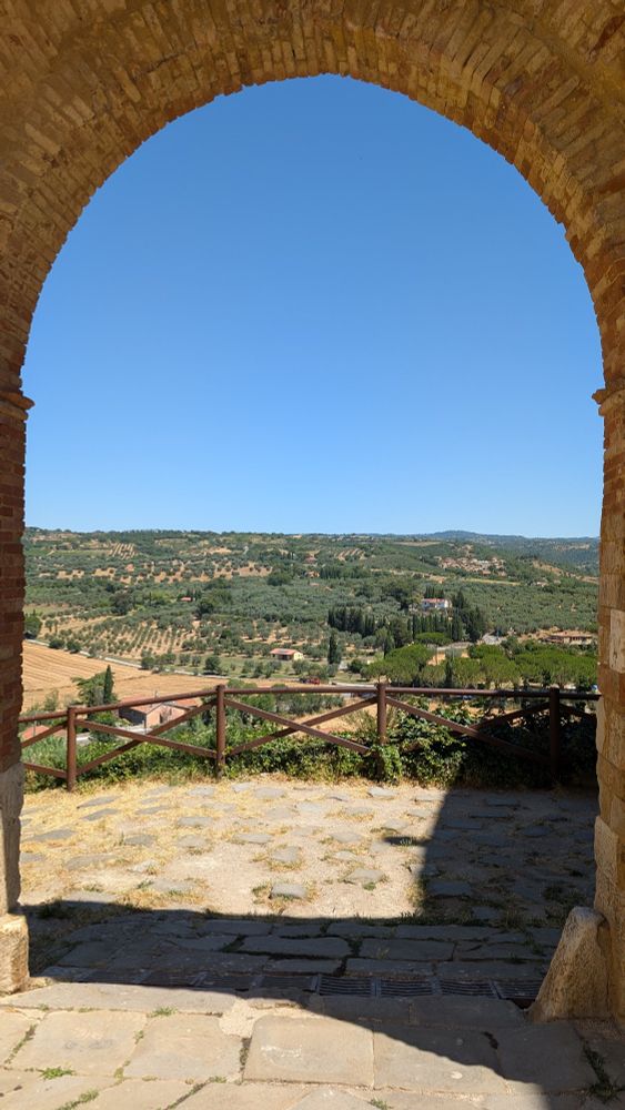 Vue à travers une arche en pierre de Magliano en Toscane, montrant un paysage vallonné avec des oliveraies, des cyprès et des champs dorés sous un ciel bleu clair. Une clôture en bois délimite le point de vue au premier plan, et la lumière du soleil projette des ombres sur le sol pavé.