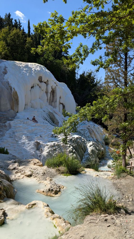 Cette photo montre les célèbres formations calcaires blanches de Bagni San Filippo en Toscane. On peut voir les terrasses naturelles de travertin blanc créées par les dépôts de carbonate de calcium des sources chaudes, qui s'élèvent en gradins sculptés par l'eau. Des bassins d'eau turquoise s'étalent à différents niveaux, alimentés par les sources thermales. La formation rocheuse blanche contraste magnifiquement avec la végétation méditerranéenne environnante - cyprès, chênes verts et autres arbres et arbustes qui encadrent le site. Le ciel bleu parsemé de quelques nuages blancs complète ce paysage naturel exceptionnel. On aperçoit une personne assise sur les formations calcaires, donnant une échelle à ces impressionnantes créations géologiques sculptées par des millénaires d'activité thermale.