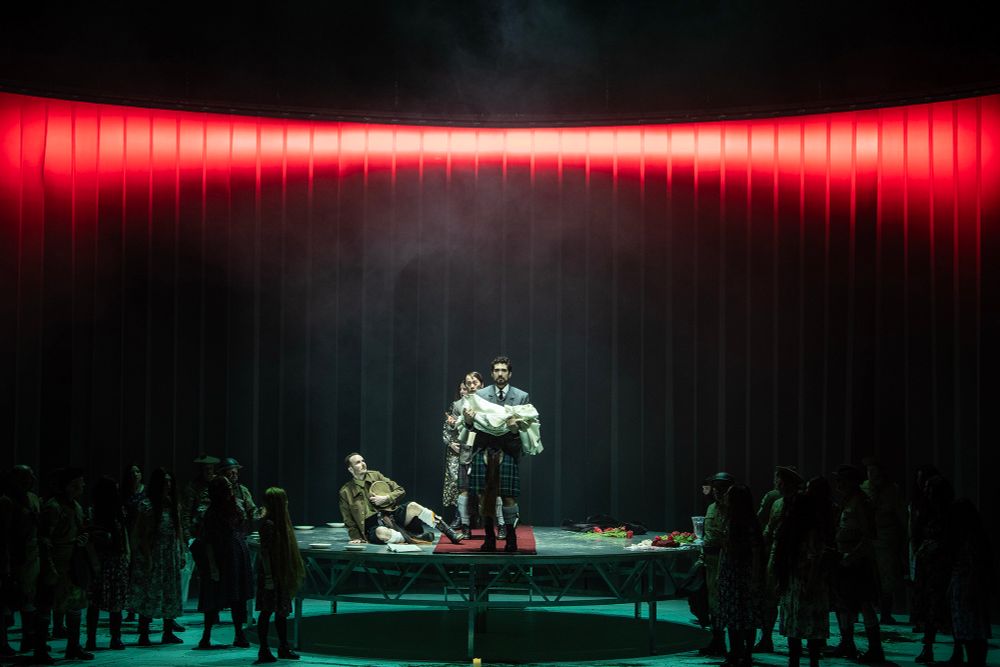 Man holds white coat in arms on stage with long, horizontal red light at top and green-hued lighting at the bottom. He wears a Scottish kilt. People are scattered on the stage in shadowy silhouettes. Another man is on the ground in shock in a green military jacket and kilt. Others stand behind man with white jacket in arms. 