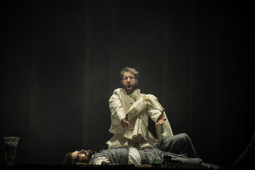 Brent Michael Smith as Banco in Macbeth lies on ground in a grey vest and pants with a white dress shirt underneath the vest, killed by assassins. Macbeth see his body in a vision and is kneeling over him singing in torment over the murderous act he hired. He is dressed in regal white with a white sash of wool over his left shoulder, and stretched out his arms, gesturing towards Banco‘s corpse. The lighting around is dark with a bright spotlight on the to figures. 