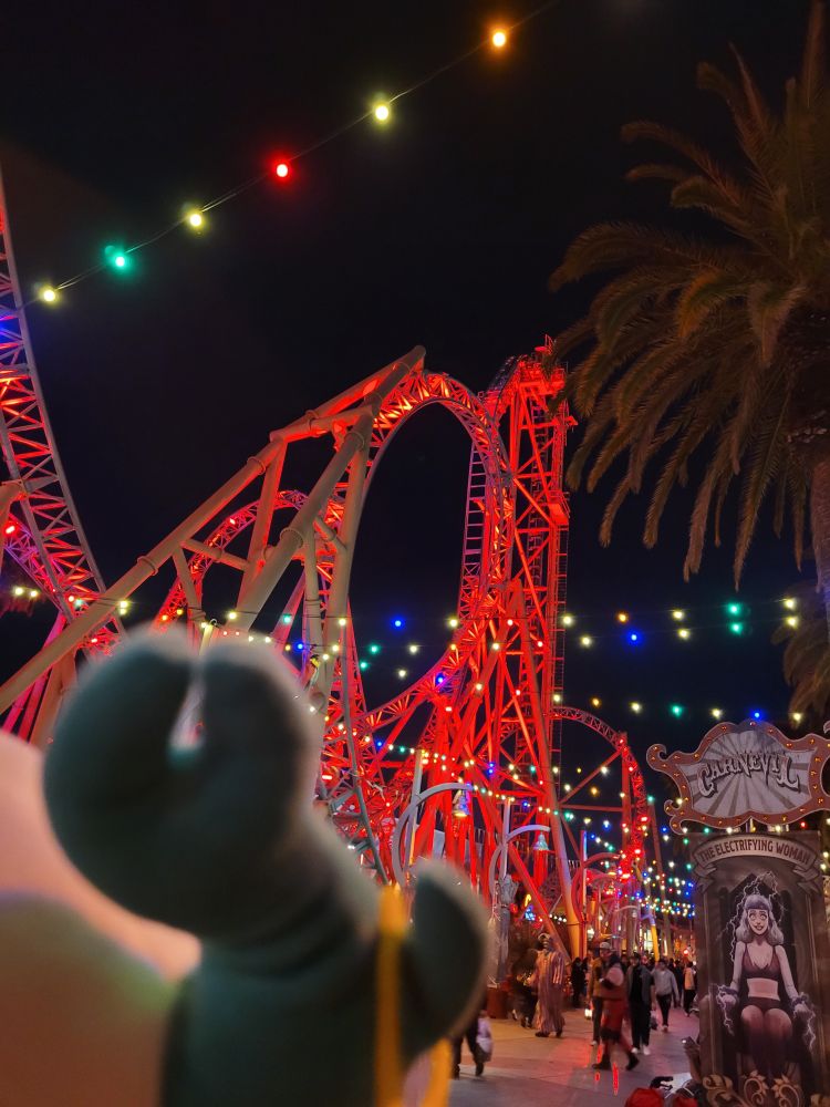 The Makeship medkit plush faces away from the camera in the bottom left corner, looking at a brightly lit roller coaster in the distance 