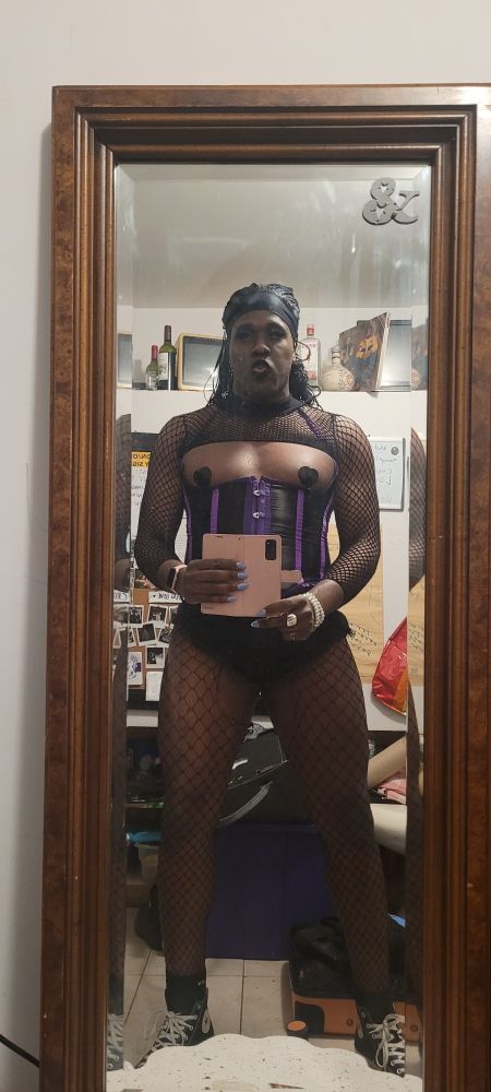 Jae Bea in a mirror selfie. Wearing an underbust corset, black jong, fishnet stockings and a fishnet shrug. 