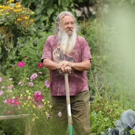 Wolf-Dieter Storl steht auf einen Spaten gestützt in seinem Garten. Er trägt eine olivfarbene Kakhi-Hose und ein burgunderfarbenes T-Shirt mit Wolfsmotiv