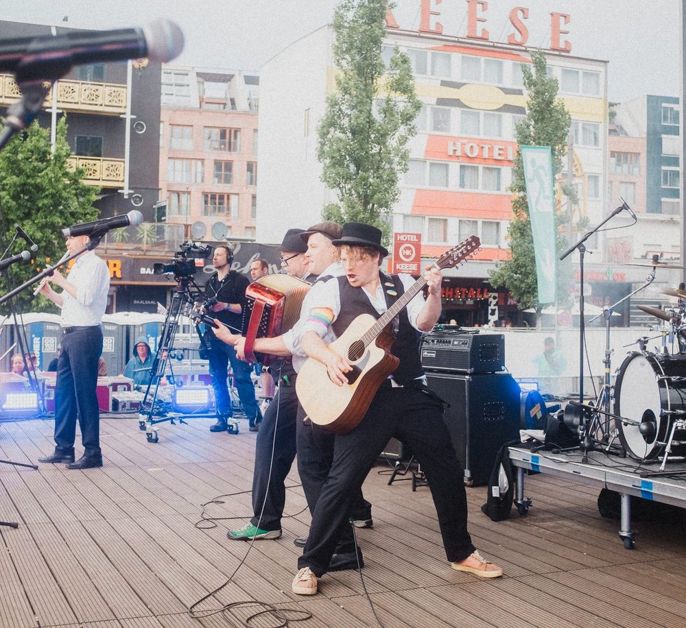 Konzertfoto der Nuthouse Flowers auf dem Spielbudenplatz in Hamburg bei der St. Pauli Aufstiegsfeier 2024.Gitarrist, Akkordeonspieler und Bassist stehen aneinander gelehnt in der Mitte der Bühne.