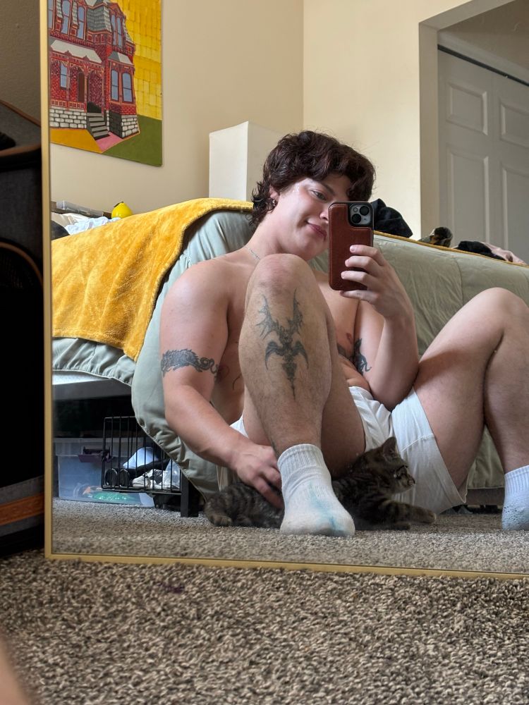 Paris sits topless at the foot of his bed playing with his little cat at the bottom of the screen. He’s wearing white shorts and white socks.