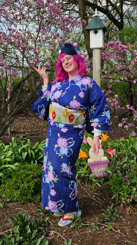 A beautiful woman with deep pink hair posing near a pink tree wearing a gorgeous deep blue yukata kimono with bright pink and blue sakura flowers print. 