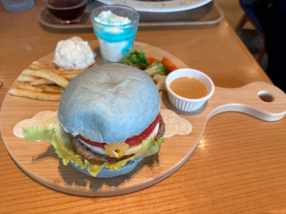 青いバンズにトマト、目玉焼き、パティ、レタスが挟まれたバーガー。トマトのところに黄色い鈴型のチーズがついており、ドラえもんカラーになっている。そのうしろにはフライドポテトやポテトサラダ、蒸し野菜、ヨーグルト（ブルーハワイソースがかかっている）が並んでいる。