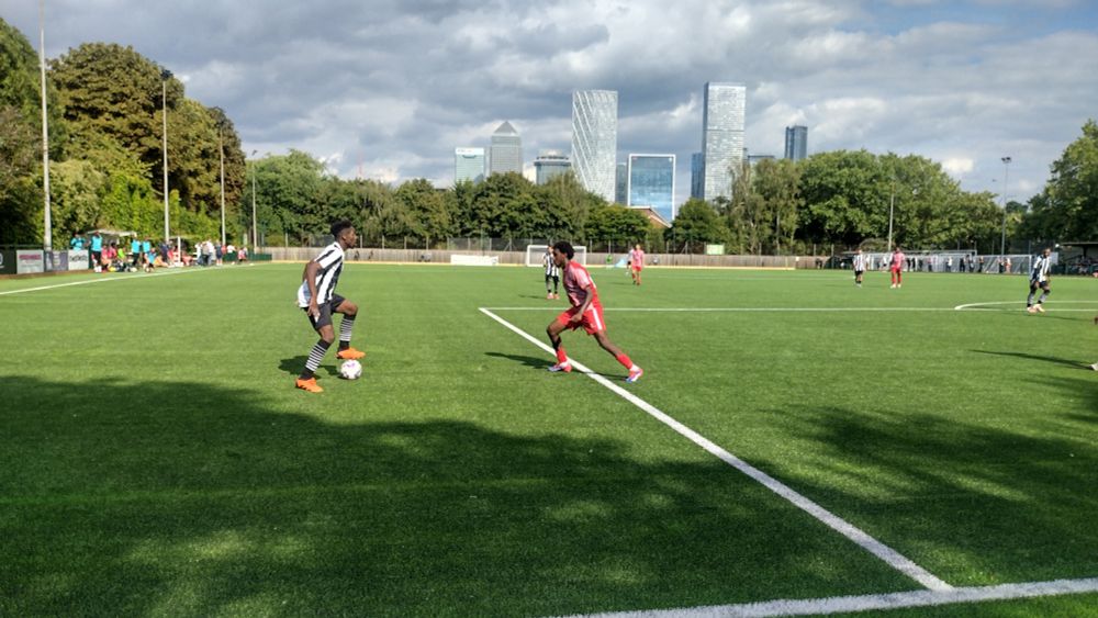 Two footballers facing off in the sunshine with the towers at canary wharf in the background
