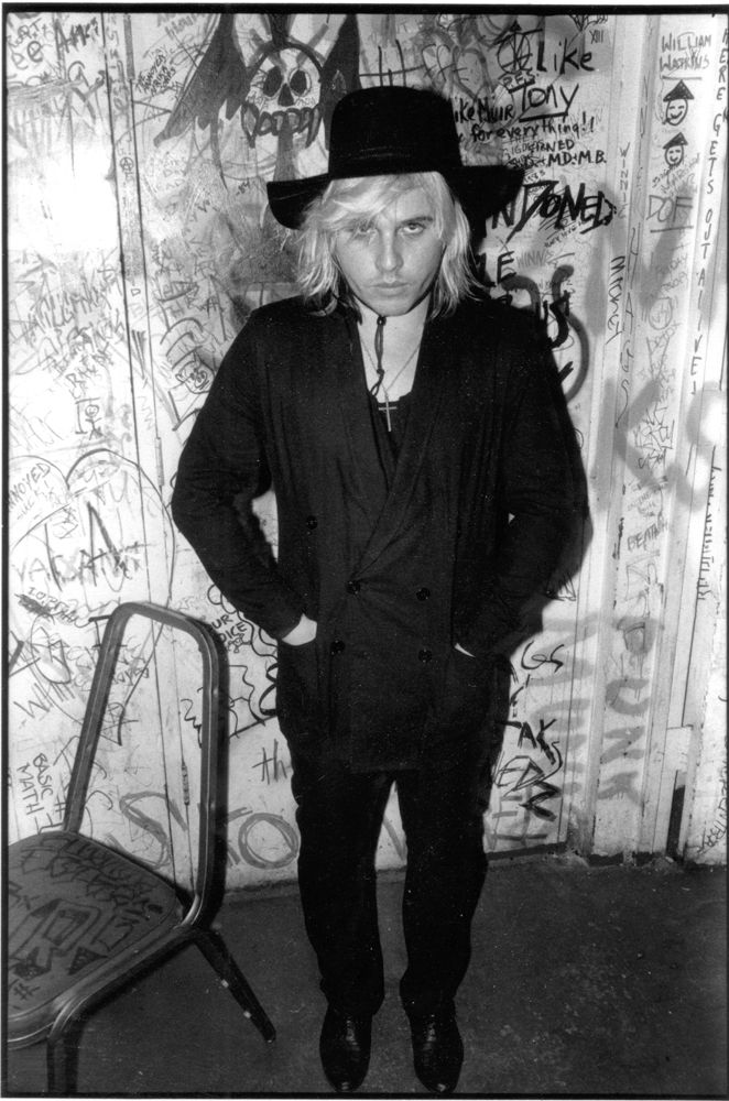 jeffrey lee pierce standing against a graffiti wall wearing all black and a cowboy hat