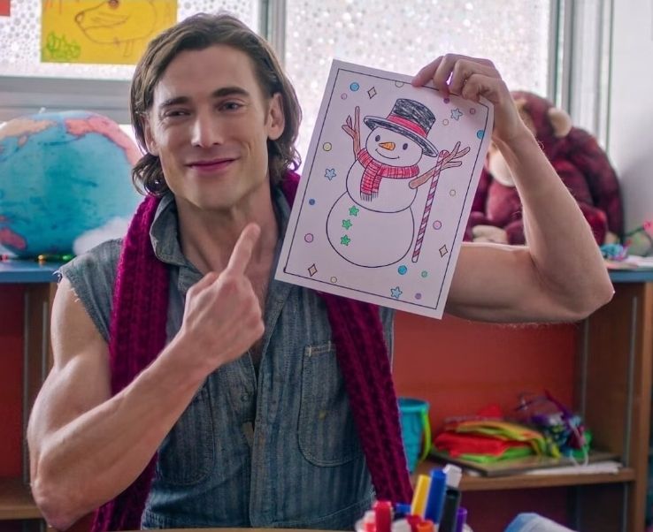 White guy wearing sleeveless jump suit and a red scarf holding up a drawing of a snowman pointing to himself. 