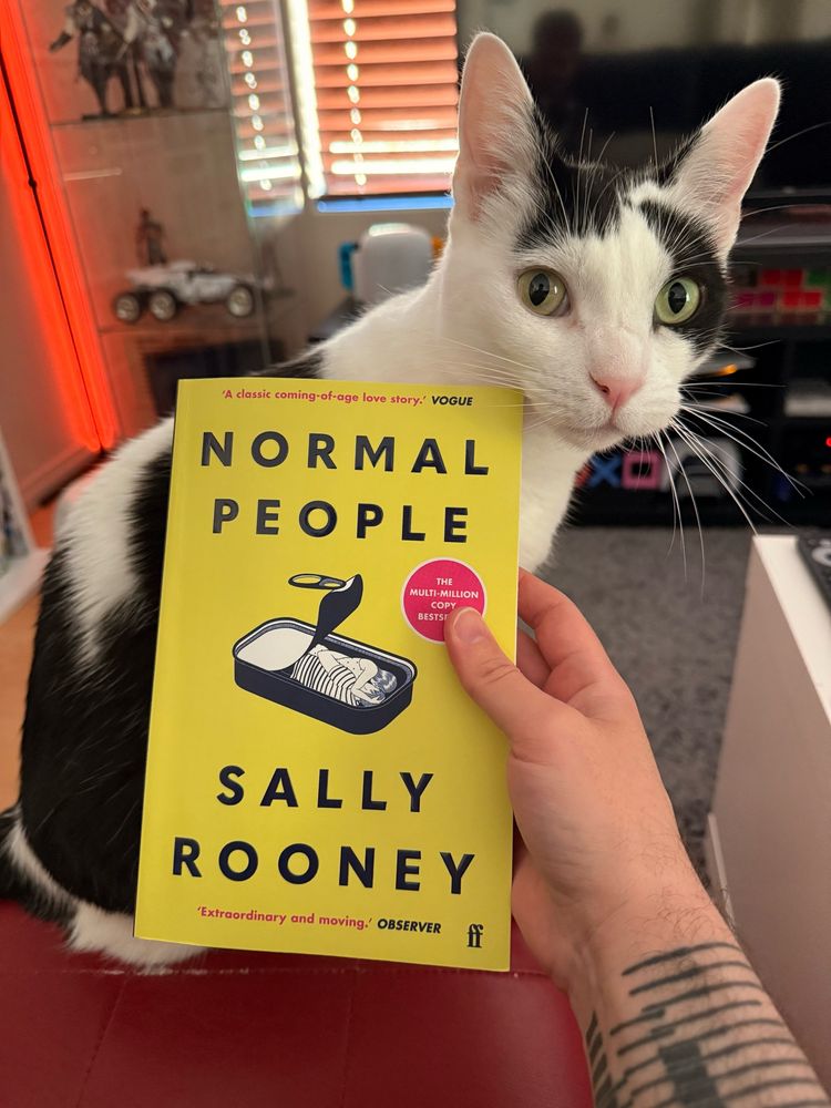 The paperback copy of Normal People by Sally Rooney held up in front of Krem, a black and white shorthair cat who is looking at the camera in a very needy way (it’s dinner time).