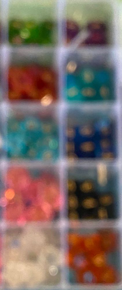 An out of focus photo of many dice grouped in 10 different colors (green, purple, red, teal, light blue, dark blue, pink, black, white, orange) inked in gold.