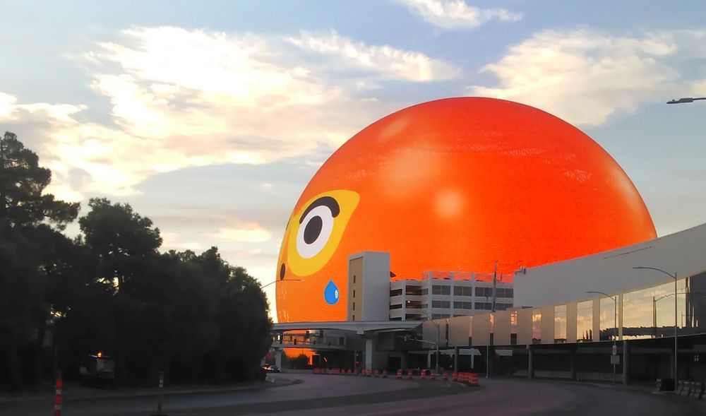 The Las Vegas Sphere (whose name is Orbi, FYI) showing a happy face, but instead of being yellow, she's orange from a "sunburn," except around the eyes, where she just took off her sunglasses.  There is also a little drop of sweat running down her face. Don't forget your sunscreen people!