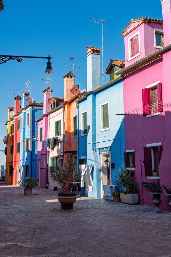 A row of brightly coloured houses in  Burano variously painted yellow, orange, blue and pink. The sky is deep blue. There is washing drying outside some of the houses. 