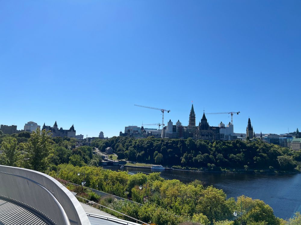 South view from Kìwekì Point at the end of August. Parliament Hill, Kichi-Sìbì (Ottawa River), the Ottawa Locks, and the Chateau Laurier are in view under a clear blue sky. All the trees are full and green.