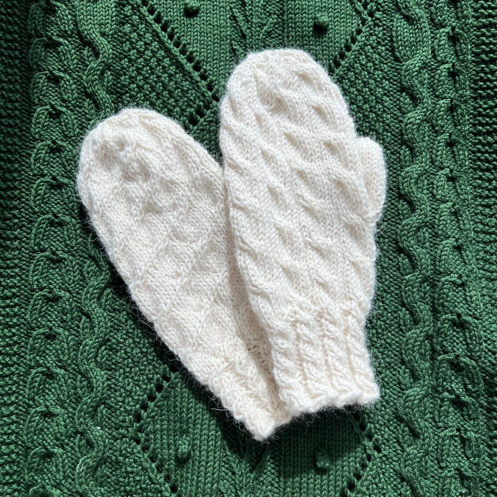 A pair of white handknit mittens against a dark green knitted lap blanket.