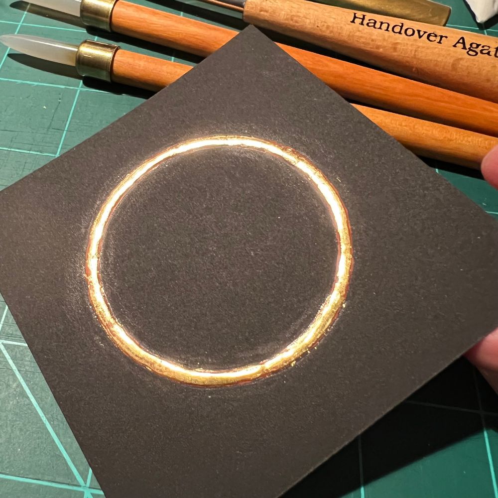 A square black piece of paper with a gilded ring of gold with agate burnishers in the background. 