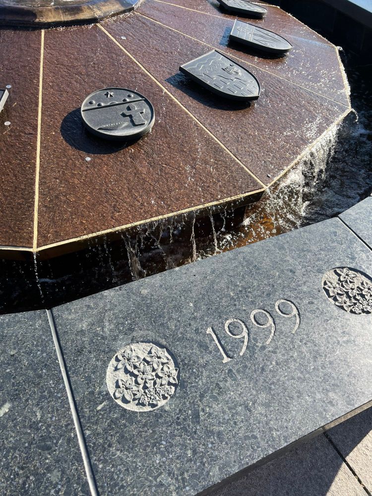 Centennial Flame in front of Parliament showing the ᓄᓇᕗᑦ (Nunavut) bronze circular shield on the fountain showing its coat of arms above with the year 1999 carved into granite ledge with the territorial flower of purple saxifrage carved into roundels flanking the year.