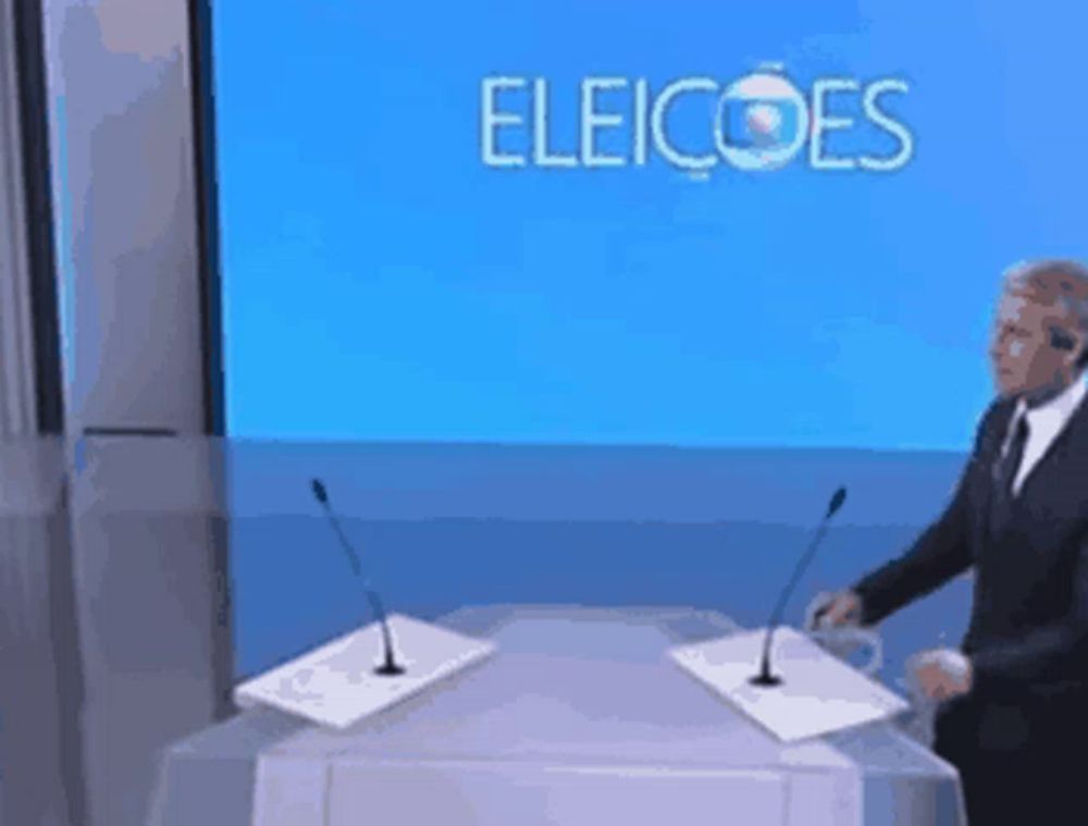 a man stands at a podium with two microphones in front of a screen that says eleicoes