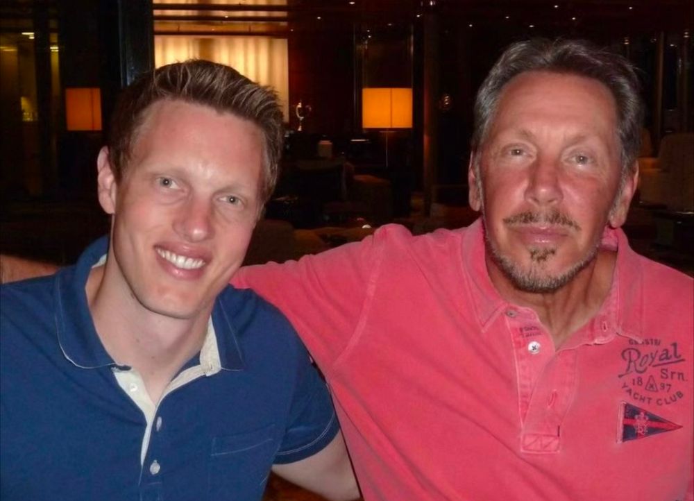 A white blond man with a giant forehead, blinding white teeth, clear plastic surgery, and lip filler wearing a blue polo shirt next to a white man in a pink polo shirt who also is white, with greying blond hair, a giant forehead, similar plastic surgery and a skimpy mustache and beard. 