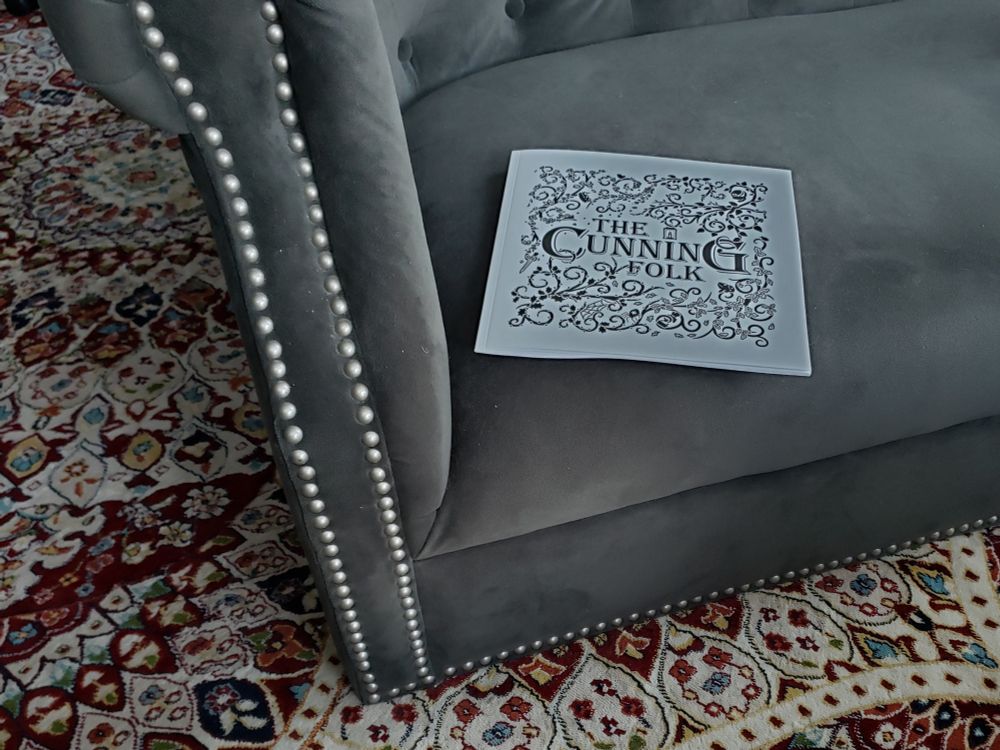 An independent TTRPG, "The Cunning Folk," sits 9n a sofa cushion. The blue/gray sofa is missing it's legs and set flush on a patterned rug.