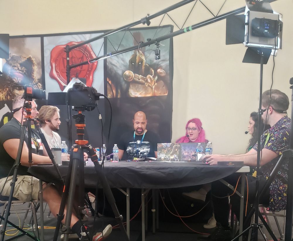 A group of people playing the TTRPG "Shadowrun: Anarchy" (a fast and loose version of the notoriously rules-heavy cyberpunk TTRPG) on a stage at Gen Con.

The image is focused on two people, B. Dave Walters (a popular Actual Play actor) and Rem Alternis (media manager for Catalyst Game Labs).