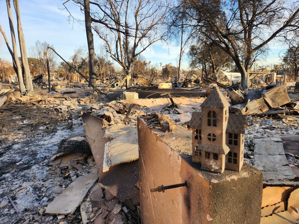 Burned remains of house with metal Christmas house decoration on wall in front.