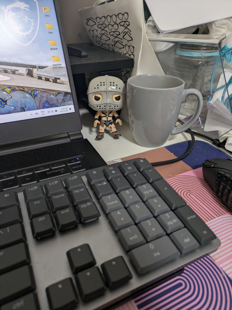 A picture of the author's laptop and keyboard, tea in a grey cup, a Funko Pop figurine of The Humungus from Mad Max 2 aka The Road Warrior, and accumulated clutter on the desk behind