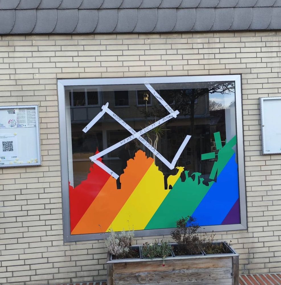 Es ist ein Fenster des queeren Zentrums "Spektrum" in Gifhorn zu sehen. Silhouetten in Regenbogenfarben zeigen Sehenswürdigkeiten wie das Schloss, Ziegen und Windmühlen. Über diesen Silhouetten ist aus weißem Klebeband ein rechtsdrehendes Hakenkreuz von Angreifer*innen auf das Zentrum angebracht worden. Es ist mit Parolen beschrieben, die auf diesem Bild jedoch nicht lesbar sind.