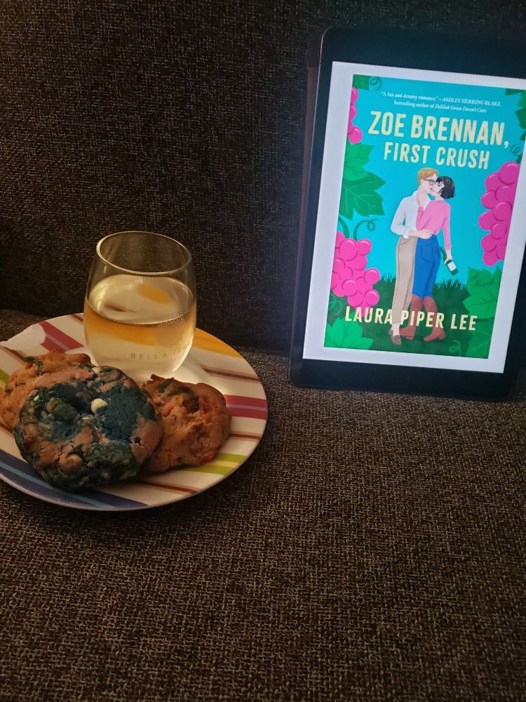 A plate of three cookies, one is pink and blue and two others have rainbow candy pieces in them. A stemless wine glass with white wine also sits in the plate. Beside it an iPad is displaying the book  Zoe Brennan, First Crush by Laura Piper Lee.