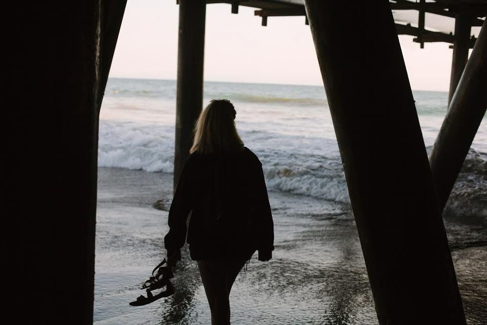 Person standing underneath an ocean pier on the shore of a beach 
