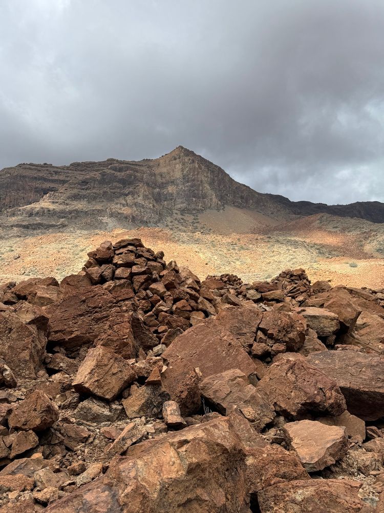 Paisaje árido con un amontonamiento de piedra roja en primer plano y una montaña más grisácea al fondo. El amontonamiento es una necrópolis grancanaria.