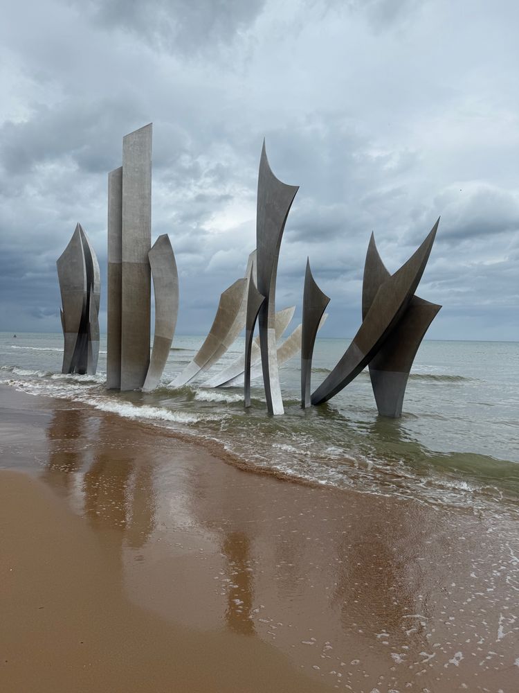 Das Foto zeigt eine moderne Skulptur am Strand, bestehend aus mehreren hohen, spitz zulaufenden Metallplatten, die aus dem Sand und dem flachen Wasser aufragen. Die Formen erinnern an Segel oder Flügel, die sich dynamisch gen Himmel strecken. Im Hintergrund ist das ruhige Meer unter einem wolkigen Himmel zu sehen, während sanfte Wellen den nassen Sand am Ufer berühren. 