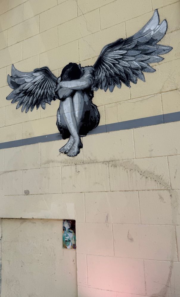 Eine Hauswand in Lille. Auf einer Linie sitzt ein Engel mit ausgebreiteten Flügeln, die Beine hochgezogen und beide Arme vor dem Kopf. Im unteren Viertel des Fotos: ein kleines quadratisches Bild einer lachenden Frau. 