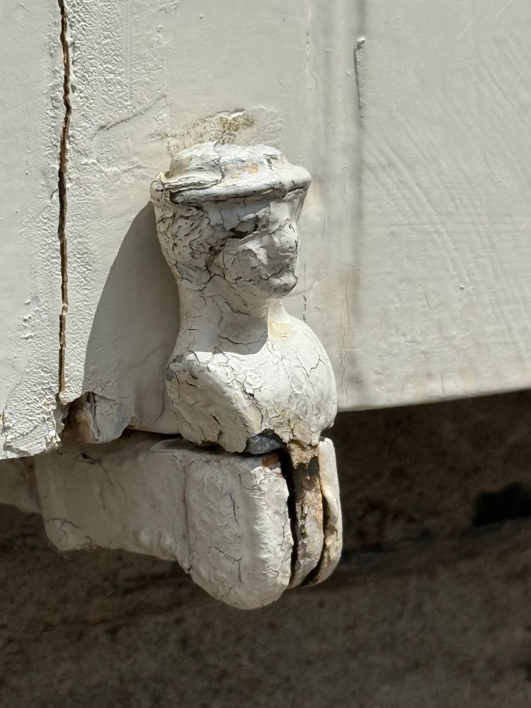 Das Foto zeigt ein altes, stark verwittertes Scharnier an einem Fensterladen. Das Scharnier ist kunstvoll geformt und stellt eine kleine menschliche Figur dar, die an der Wand befestigt ist. Die Figur scheint eine Mütze oder einen Helm zu tragen und ist dick mit weißer, rissiger Farbe überzogen, die teilweise abgeplatzt ist. Der Hintergrund besteht aus hellem, ebenfalls verwittertem Holz und einem dunkleren Stein oder Beton darunter. Das Licht fällt von oben auf die Szene und betont die Struktur der abgeplatzten Farbe und die Form der Figur.